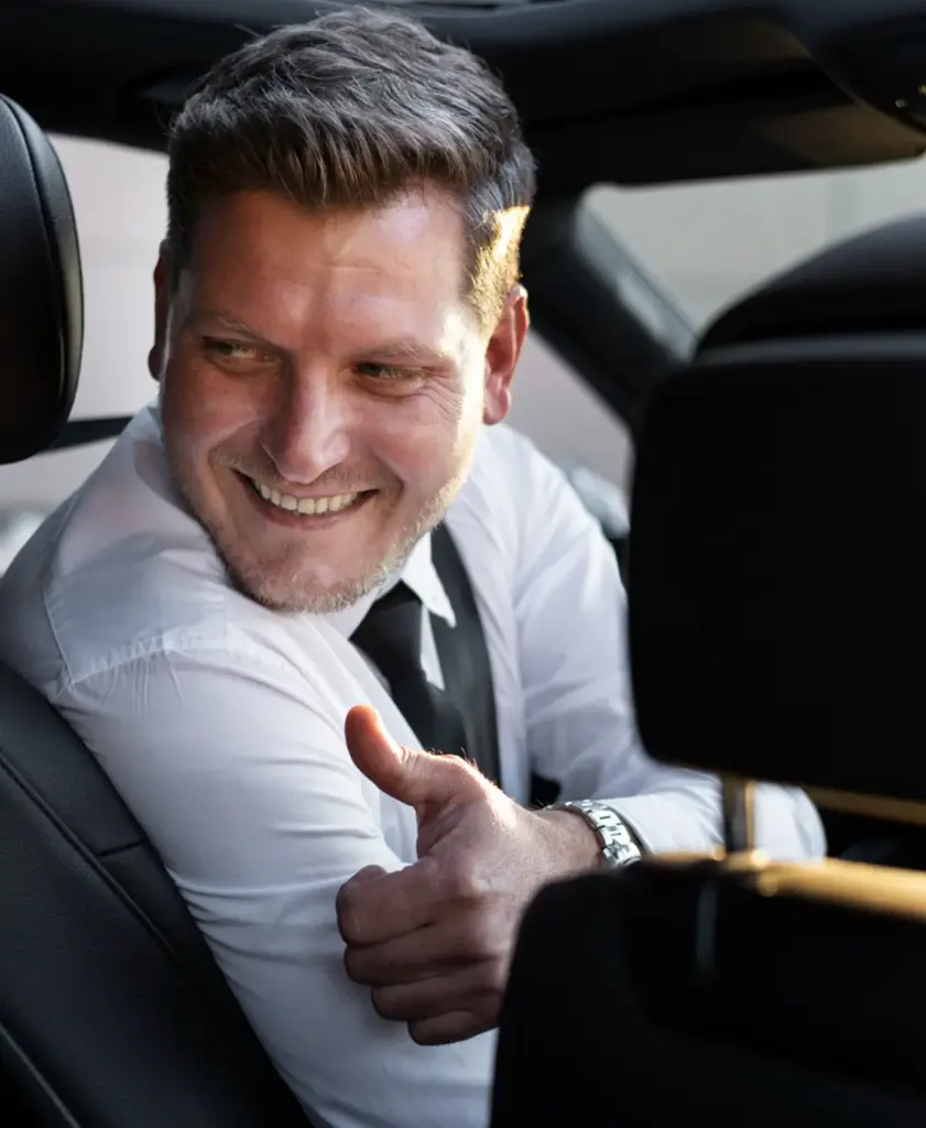 Een glimlachende chauffeur in een wit overhemd en stropdas kijkt achterom vanuit de bestuurdersstoel en steekt zijn duim omhoog, met een gedeeltelijk zichtbaar geel telefoonnummer rechtsboven
