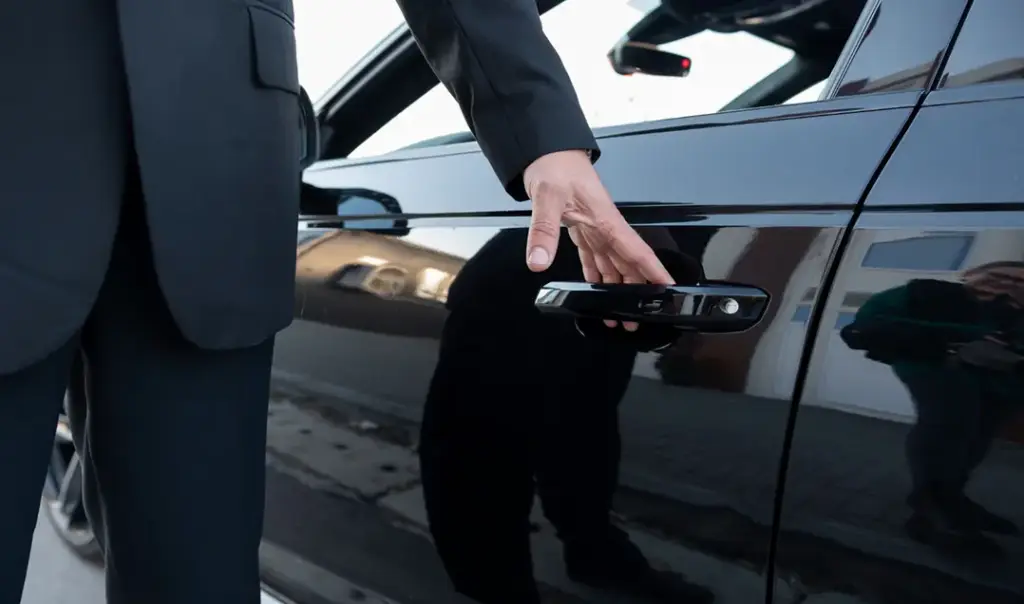 Een close-up van de hand van een chauffeur in een pak die de deurklink van een glanzend zwarte auto vastpakt om deze te openen