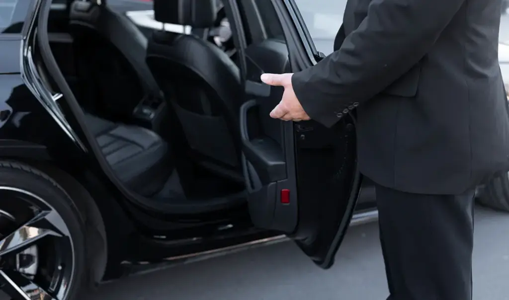 Een chauffeur in een zwart pak houdt de achterdeur van een luxe auto open, met een uitnodigend handgebaar naar het lederen interieur