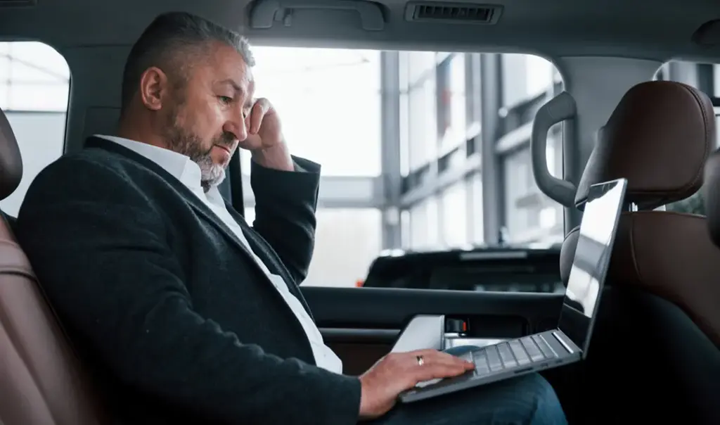 Een oudere zakenman in een jasje zit op de achterbank van een auto en werkt geconcentreerd op zijn laptop